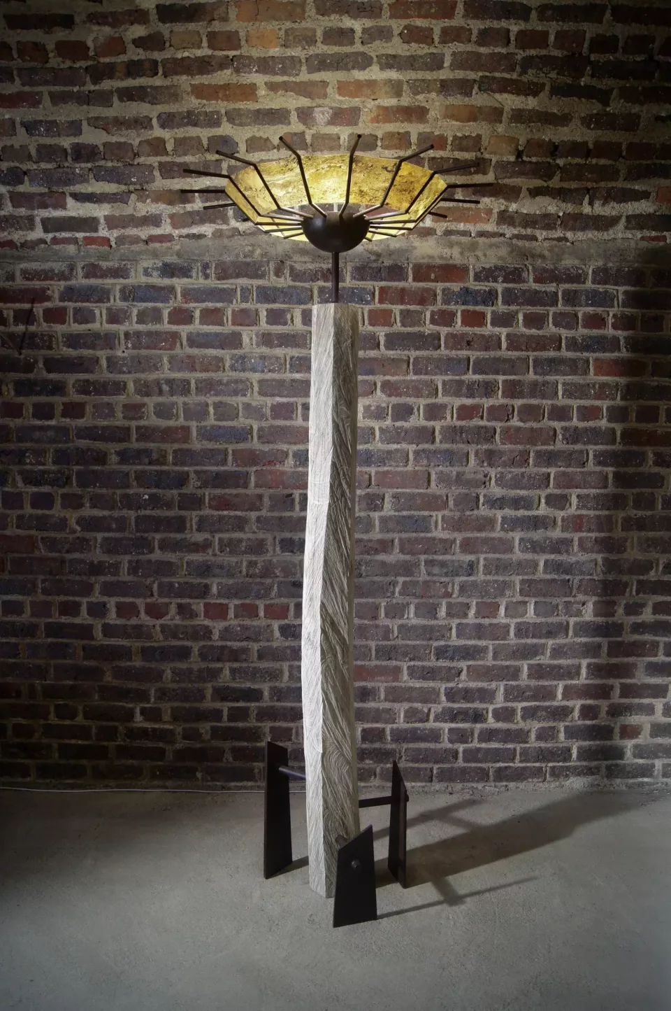 Lampadaire de deux mètres devant un mur de briques : fût de bois tronçonné, large abat-jour en éventail rayonnant.