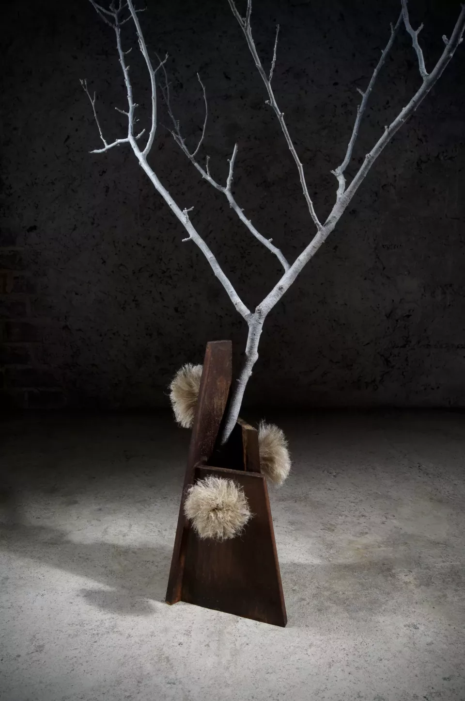 Vase trapézoïdal en acier massif rouillé, garni d’une branche blanche et de pompons de chanvre.